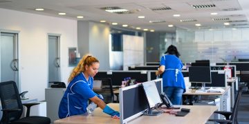 Two janitorial workers cleaning desks in open office space